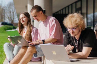 Güneşli bir günde bir grup öğrenci modern bir üniversite binasının önünde dizüstü bilgisayar çalışıyor ve kullanıyor. Yüksek kalite fotoğraf