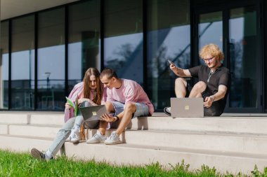 Güneşli bir günde bir grup öğrenci modern bir üniversite binasının önünde dizüstü bilgisayar çalışıyor ve kullanıyor. Yüksek kalite fotoğraf