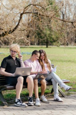 Güneşli bir günde, üç neşeli öğrenci, parkta bir bankta otururken dizüstü bilgisayar ve not defteri kullanarak birlikte çalışır ve birlikte çalışırlar. Yüksek kalite fotoğraf