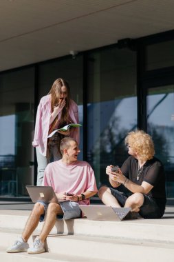 Güneşli bir günde bir grup öğrenci modern bir üniversite binasının önünde dizüstü bilgisayar çalışıyor ve kullanıyor. Yüksek kalite fotoğraf
