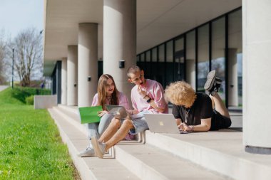 Güneşli bir günde bir grup öğrenci modern bir üniversite binasının önünde dizüstü bilgisayar çalışıyor ve kullanıyor. Yüksek kalite fotoğraf