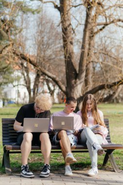 Güneşli bir günde, üç neşeli öğrenci, parkta bir bankta otururken dizüstü bilgisayar ve not defteri kullanarak birlikte çalışır ve birlikte çalışırlar. Yüksek kalite fotoğraf