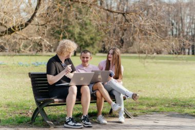 Güneşli bir günde, üç neşeli öğrenci, parkta bir bankta otururken dizüstü bilgisayar ve not defteri kullanarak birlikte çalışır ve birlikte çalışırlar. Yüksek kalite fotoğraf