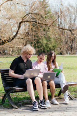 Güneşli bir günde, üç neşeli öğrenci, parkta bir bankta otururken dizüstü bilgisayar ve not defteri kullanarak birlikte çalışır ve birlikte çalışırlar. Yüksek kalite fotoğraf