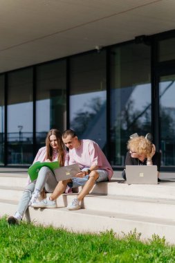 Güneşli bir günde bir grup öğrenci modern bir üniversite binasının önünde dizüstü bilgisayar çalışıyor ve kullanıyor. Yüksek kalite fotoğraf