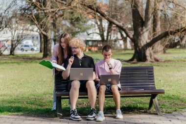 Güneşli bir günde, üç neşeli öğrenci, parkta bir bankta otururken dizüstü bilgisayar ve not defteri kullanarak birlikte çalışır ve birlikte çalışırlar. Yüksek kalite fotoğraf
