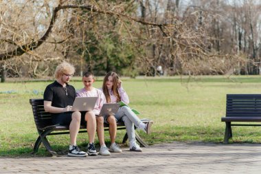 Güneşli bir günde, üç neşeli öğrenci, parkta bir bankta otururken dizüstü bilgisayar ve not defteri kullanarak birlikte çalışır ve birlikte çalışırlar. Yüksek kalite fotoğraf