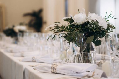 Sofistike bir kutlama için beyaz çiçekler, cam örtüler ve katlanmış peçetelerle süslenmiş zarif bir düğün yemeği masası. Yüksek kalite fotoğraf