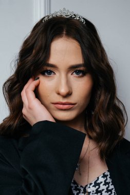 Siyah ceket ve taç giyen kendine güvenen genç bir kadın zarif beyaz bir duvara karşı çapraz kollarla duruyor, modern sofistike ve görkemli cazibeyi harmanlıyor. Yüksek kalite fotoğraf