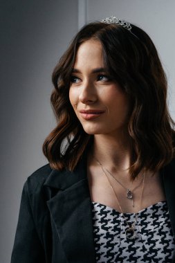 Siyah ceket ve taç giyen kendine güvenen genç bir kadın zarif beyaz bir duvara karşı çapraz kollarla duruyor, modern sofistike ve görkemli cazibeyi harmanlıyor. Yüksek kalite fotoğraf