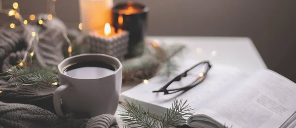 Banner image. Cozy home still life concept. Cup of hot coffee and opened book with atmospheric lights and burning candle. Winter holidays, Christmas time concept. Long banner for web site.