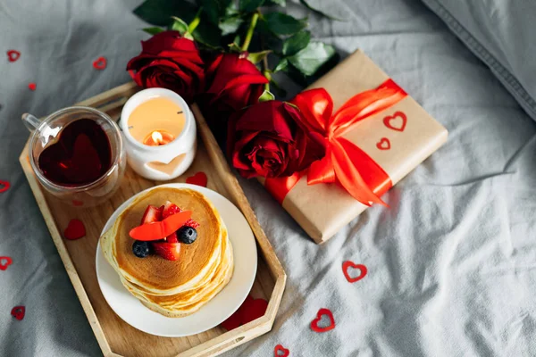 Valentine\'s day breakfast concept. Pancakes with berries, roses flowers, cup of tea, gift box and candle in candlestick. Top view.