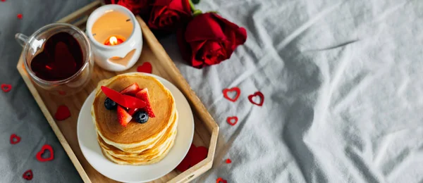 Pancakes in the shape of a heart with berries, roses flowers, cup of tea and candle in candlestick. Valentine\'s day breakfast concept. Top view with copy space. Banner for design, web site.