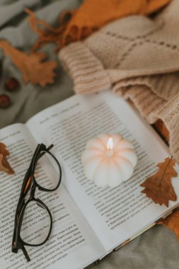 Beyaz yanan mum şeklinde balkabağı açık kitap, gözlük, rahat kazak ve yatakta sonbahar yaprakları.