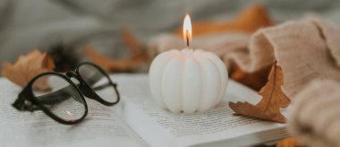Açık kitap üzerinde yanan balkabağı şeklinde beyaz mum, gözlük, rahat süveter ve yatakta sonbahar yaprakları. Tasarım için uzun pankart, web sitesi