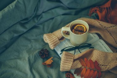 Açık kitaplı koyu renkli atmosferik fotoğraf, bir fincan limon çayı, sıcak süveter, gözlük ve gri yatakta sonbahar yaprakları. Boşluğu kopyala