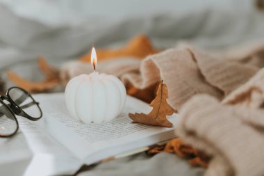 Beyaz yanan mum şeklinde balkabağı açık kitap, gözlük, rahat kazak ve yatakta sonbahar yaprakları.