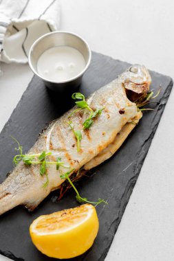 Baharatlı ızgara balık deniz kabuğu, yeşil limon ve siyah tahta tabakta ışık masasında mikro yeşil. Üst görünüm.