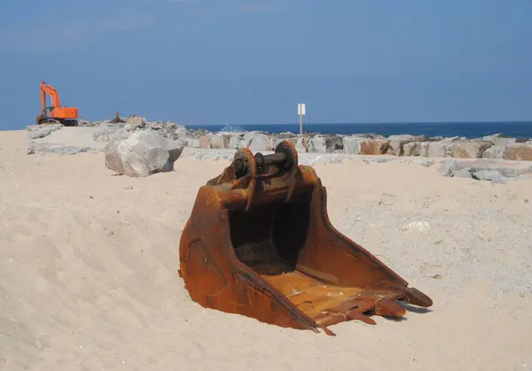 Kumsalda kazıcı bağlantısı, paslı eski kazı kovası, kirlilik, çevresel yıkım.