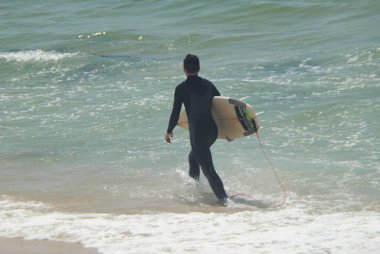 Sahilde sörfçü kolunun altında sörf tahtasıyla deniz suyuna giriyor.