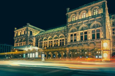 The vienna state opera house shines brightly at night. Casting a golden glow as cars and buses zoom by. Leaving light trails in their wake clipart