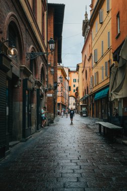 Turistler ve vatandaşlar, yağmurdan sonra İtalya 'nın Bologna şehrinin merkezinde renkli binalar ve sokak lambalarıyla karakteristik bir ıslak kaldırım taşı caddesi boyunca yürüyorlar.