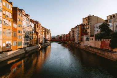 Onyar nehri kırmızı bir köprünün altında huzurlu bir şekilde akar, Girona, katalonya 'daki geleneksel binaların sıcak tonlarını yansıtarak, gün batımında resim gibi bir manzara yaratır.