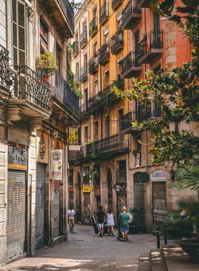 Bagaj taşıyan ve bebek arabalarını iten turistler, Barselona 'nın gotik bölgesinde renkli binalar ve işlenmiş demir balkonlarla dolu dar bir sokakta yürüyorlar.