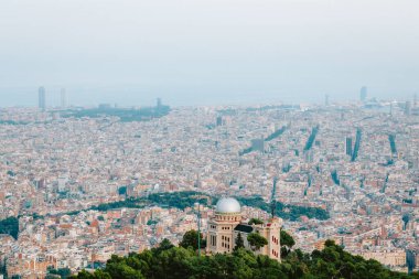 Fabra rasathanesi ve ön planda Tibidabo tepesindeki ağaçların etrafındaki deniz ve sagrada ailesi de dahil olmak üzere Barselona şehri sisli bir günde uzak mesafelere uzanır.