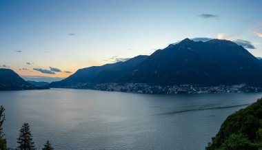 Sıcak günbatımı renkleri göl kenarına gökyüzünü boyar sakin sulara, büyüleyici köylere ve görkemli dağlara yansıyarak huzur verici bir manzara yaratır.