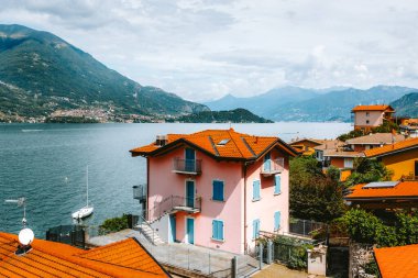 Lezzeno 'da pembe bir evin manzarası turuncu kiremitlerle kaplı göl kenarlarında bir yelkenli, uzak dağlar ve huzurlu bir atmosferi var.