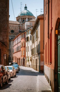 Parma Katedral Kubbesi, güneşli bir yaz gününde, parka parkedilmiş arabaların ve renkli binaların bulunduğu dar bir caddenin üzerinde yükseliyor.