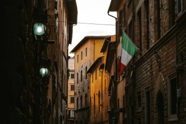 Dar bir Floransa caddesinde, gün batımını aydınlatan sıcak bir gün batımı ışığı. Binaya asılı İtalyan bayrağı ve klasik sokak lambaları büyüleyici atmosfere yeni bir hava katıyor.