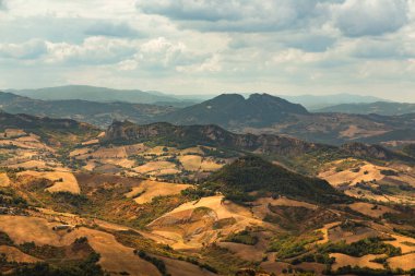 Güneş ışığı, San Marino 'nun pitoresk kırsalındaki altın tarlaları ve ağaçlık tepeleri aydınlatır. Bulutlu bir gökyüzünün altında sakin bir manzara yaratır.