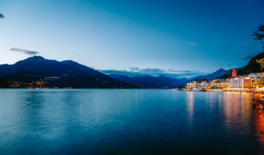 Lombardy, İtalya 'daki çarpıcı bir dağ silsilesinin çerçevelediği Como Gölü üzerindeki Bellagio köyünün büyüleyici ışıklarını yansıtan sakin sular.