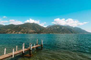Como Gölü 'nde sakin bir manzara, sakin sulara uzanan yıpranmış bir ahşap iskele, büyüleyici Menaggio köyü ve güneşli mavi gökyüzünün altında yemyeşil dağlar.
