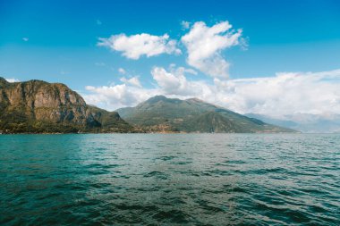 Lombardy, İtalya 'daki Como Gölü, beyaz bulutlarla benekli canlı mavi gökyüzünün altından yükselen dağlarla nefes kesici bir manzara sunar.