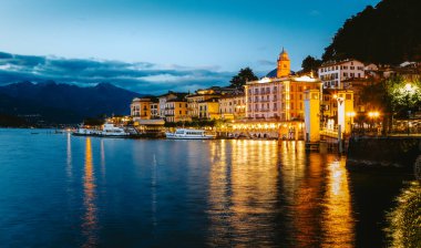 Como Gölü, Bellagio köyünün sıcak ışıklarını ve alacakaranlıktaki tekneleri yansıtıyor. Dağlar ve bulutlu gökyüzü de arka planda, Lombardy, İtalya 'da.