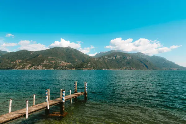 Como Gölü 'nde sakin bir manzara, sakin sulara uzanan yıpranmış bir ahşap iskele, büyüleyici Menaggio köyü ve güneşli mavi gökyüzünün altında yemyeşil dağlar.
