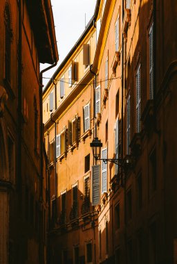 Sıcak güneş ışığı, İtalya 'nın Verona kentinde beyaz panjurların ve demir balkonların yer aldığı geleneksel binalarla dolu dar bir sokağı aydınlatarak İtalyan kentsel yaşamının pitoresk bir manzarasını yaratıyor.