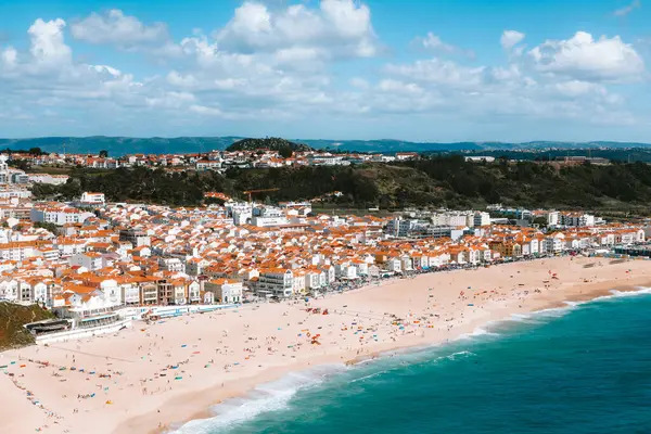 Nazare plajını ve kasabasını kaplayan hava manzarası tüylü bulutlarla bezenmiş çarpıcı mavi gökyüzünün altında, güzel Portekiz kıyıları boyunca yaz tatillerinin tadını çıkaran turistleri vurguluyor.