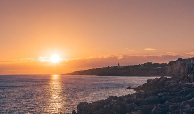 Güneş Atlantik Okyanusu 'nun üzerinde batıyor, Cascais' in kayalık kıyı şeridine sıcak bir parıltı saçıyor, Santa Marta deniz feneri uzakta dik duruyor.