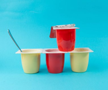 Yoğurt bardakları seti, plastik kaplarda pembe ve sarı meyveli yoğurt, kaşıklı süt pudingi kabı, renkli arka planda küçük bir paket süt ürünü.