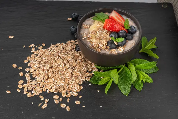 Siyah kasede pişirilmiş. Muzlu yulaf ezmesi sütlü yulaf lapası, çilek ve yabanmersini haşlanmış emmer, sağlıklı tahıl gevreği. Koyu arkaplanda taze yeşil naneli buğday taneleri.