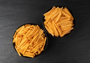 Kızarmış cips çubukları siyah kaselerde, parmak patates cipsleri, altın ayakkabı teli patates kızartmaları, patates kızartmaları, kızarmış patatesler, kızarmış patatesler, koyu taştan masa arkasında.