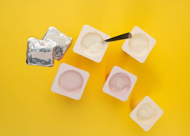 Yoğurt bardakları seti, plastik kaplarda pembe ve sarı meyveli yoğurt, kaşıklı süt pudingi kabı, renkli arka planda küçük bir paket süt ürünü.