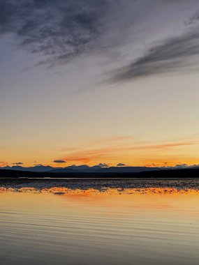Bir gölün sakin yüzeyi, uzaktaki barışçıl bir dağ sırası tarafından çerçevelenmiş, canlı günbatımı gökyüzünü yansıtır..