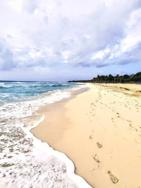 Uzakta köpüklü okyanus dalgaları ve palmiye ağaçları olan kumlu bir sahil boyunca ayak izleri var..