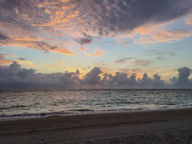Dağınık bulutlar ve altın renkleriyle okyanusun üzerinde dramatik bir gün batımı. Ön plandaki kumlu plaj kompozisyona doku ve derinlik ekler..