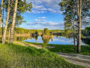 Gün batımında etrafındaki ağaçları ve parçalı bulutlu gökyüzünü yansıtan huzurlu bir göl. Kıyı şeridi boyunca yemyeşil alan manzaralı açık hava güzelliğini artırıyor..
